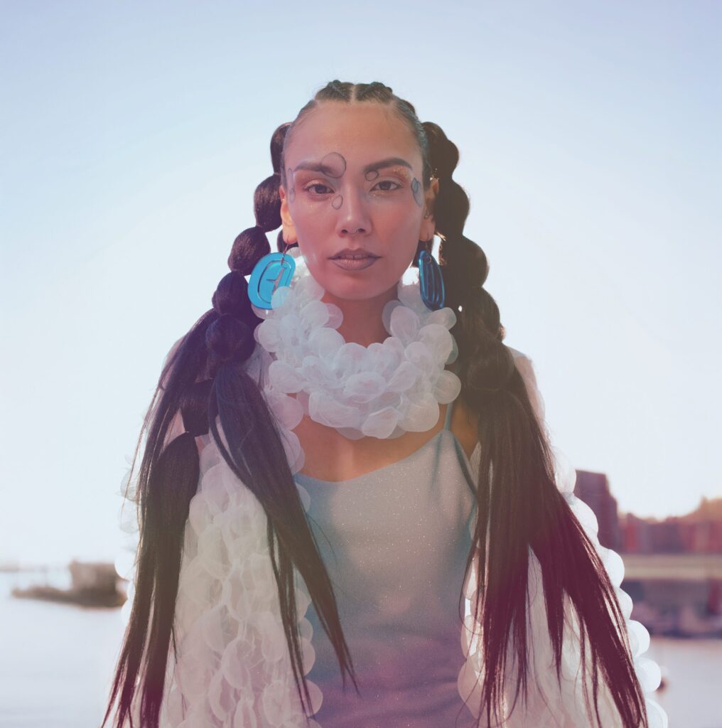 A Vancouver Indigenous Fashion Week model with long, dark, braided hair looking forward in a light blue dress and wearing a light bubbly scarf
